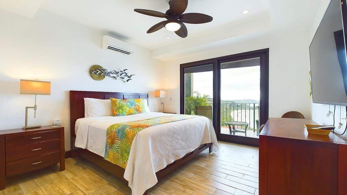 A hotel room with a bed, a ceiling fan, a TV, a balcony with a view, and a wooden floor.