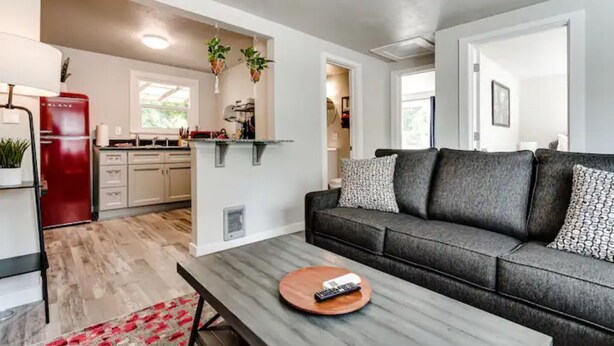 A modern living room with a grey sofa, a coffee table with a remote control, and a kitchen area with a red refrigerator and hanging plants. - Remodeled 2 bdrm duplex near the U of O (Eugene)