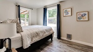 A bedroom with a bed, a bedside table, a lamp, a window with curtains, and two wall art pieces. - Remodeled 2 bdrm duplex near the U of O (Eugene)