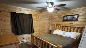 Une chambre dans une cabane en bois, avec un lit, un ventilateur de plafond, une fenêtre avec des rideaux et un petit bureau.