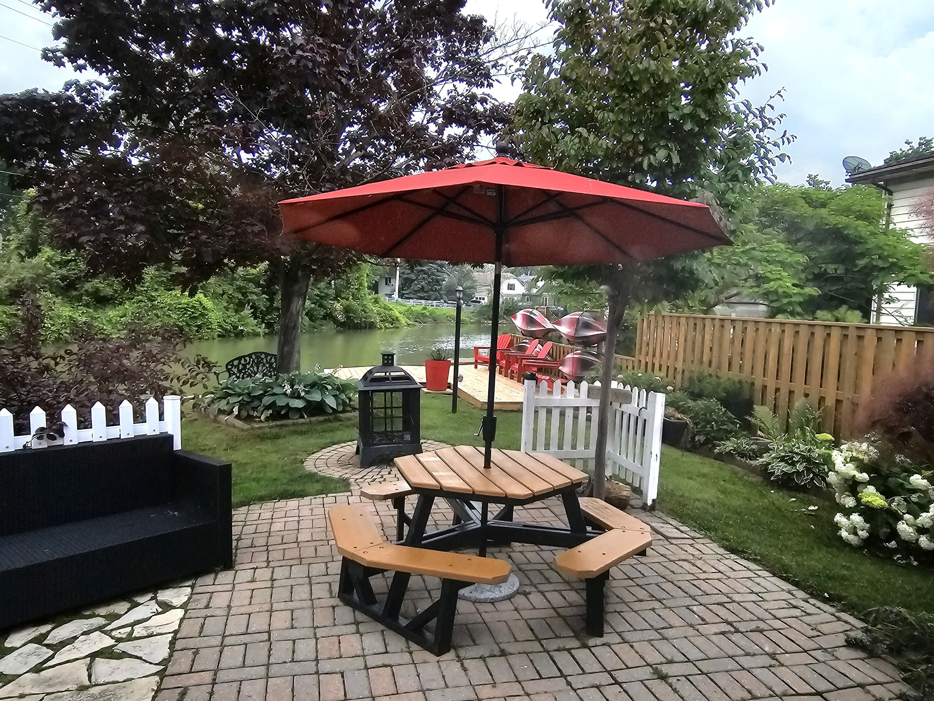 A patio with a picnic table and umbrella, surrounded by a wooden fence, a white picket fence, and a garden with various plants and a pond.
