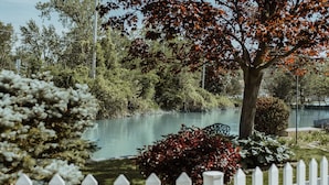 A serene lakeside view with a white picket fence, lush greenery, and a calm body of water.