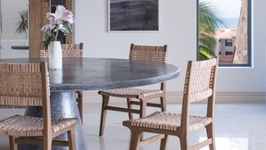 Un comedor con una mesa redonda, sillas de mimbre, un jarrón con flores, un cuadro enmarcado y un amplio ventanal con vistas.