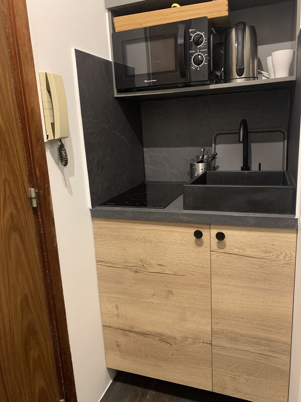 Een kleine keuken met een magnetron, broodrooster en koffiezetapparaat op een zwart aanrecht. Er is een spoelbak met een kraan en een houten kastje eronder.