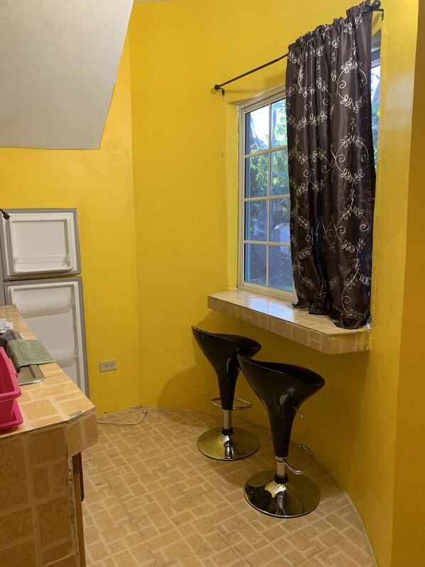 Une petite salle de bain aux murs jaunes, une fenêtre avec un rideau, deux tabourets de bar noirs et un lavabo blanc.