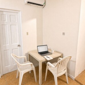 Une petite pièce avec une porte blanche, un ordinateur portable posé sur une table, deux chaises blanches et un appareil de climatisation fixé au mur.