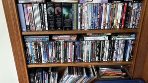Ein Bücherregal mit DVDs und Büchern, eine Holz-Uhr und eine blaue Aufbewahrungsbox.