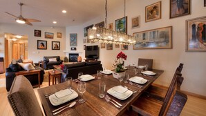 Un espace repas avec une longue table en bois, dressée pour un repas, entourée de chaises et d’un canapé.