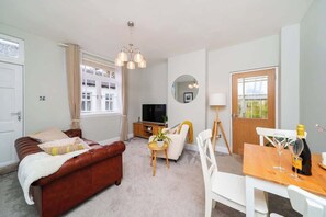 A furnished living room with a brown sofa, a dining table with two chairs, a TV, and a small plant.