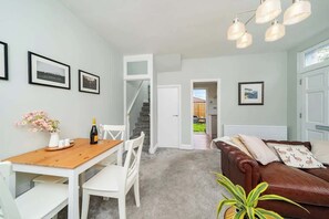 A room with a wooden dining table, white chairs, a brown leather sofa, and a staircase leading to another area.