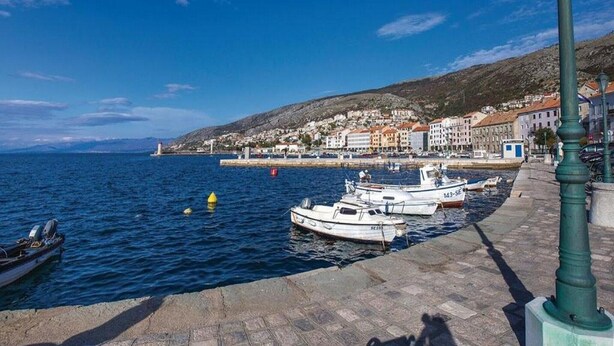 Un pueblo costero con un paseo marítimo empedrado, barcos amarrados a lo largo del frente marítimo y edificios con tejados de tejas rojas.