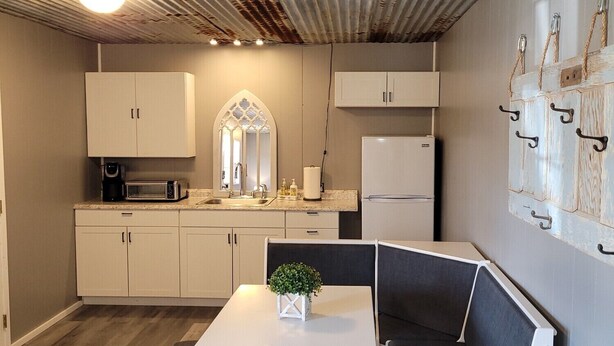 A compact kitchen with white cabinets, a microwave, and a refrigerator. There's a small table with a plant and a bench. - Safari-ish Cabin Getaway in Jacksboro (Jacksboro)