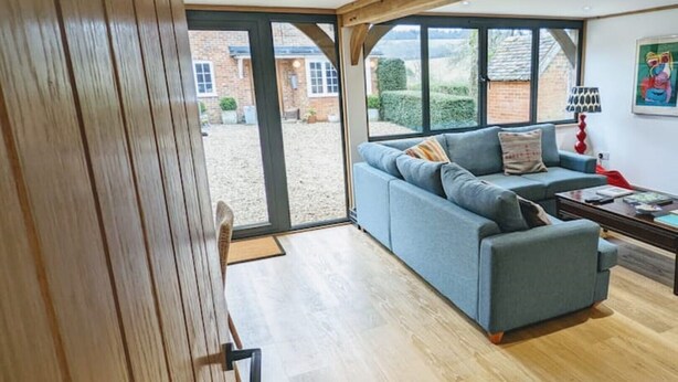 Ein modernes Wohnzimmer mit einem blauen Sofa, Holzboden und großen Fenstern mit Blick auf ein Ziegelgebäude und Grünflächen.