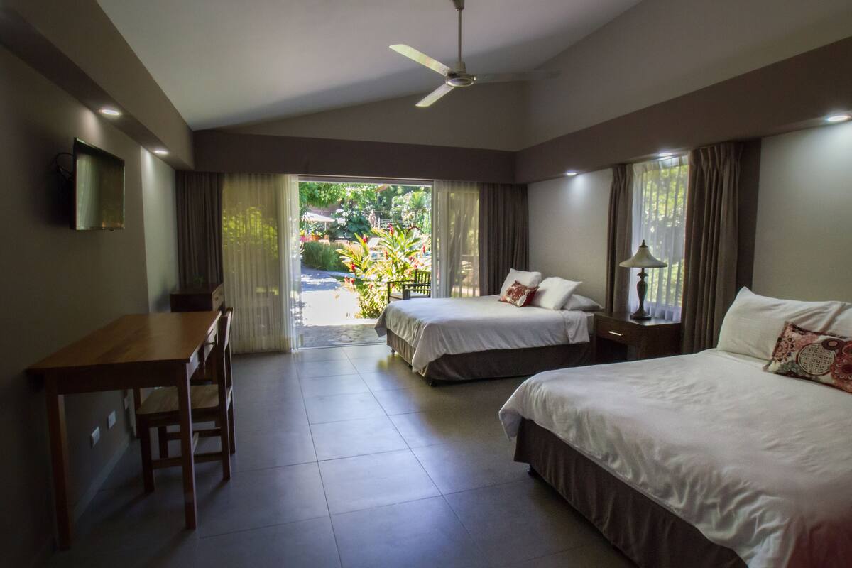 A hotel room with two beds, a desk, and a chair. There is a ceiling fan, a lamp, and a view of the outdoors.