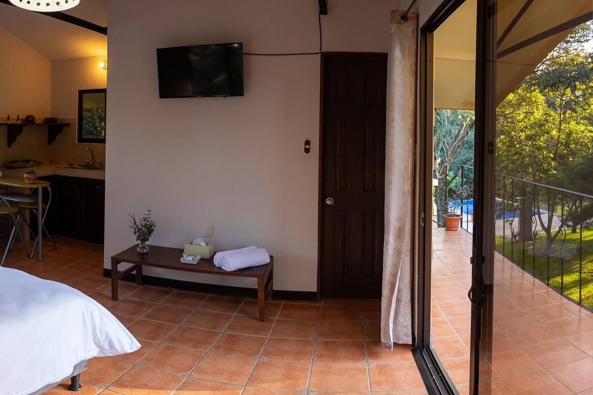 A bedroom with a bed, a red sofa, a small table with a vase, and a balcony with a view of trees.