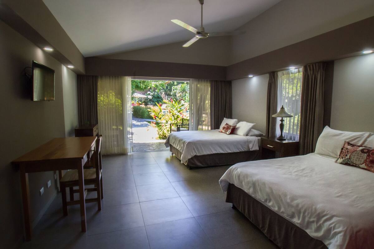 A hotel room with two beds, a desk, and a chair. There is a ceiling fan, a lamp, and a view of the outdoors.