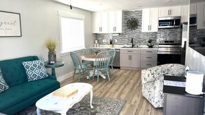 Una cocina moderna con armarios blancos, salpicadero de piedra y electrodomésticos de acero inoxidable. Cuenta con una zona de comedor con una mesa redonda y sillas, y una sala de estar con un sofá verde y un sillón estampado.