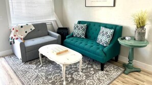 Una sala de estar con un sofá gris, un sofá azul verdoso con almohadones estampados, una mesa de centro blanca, un tapete con diseño, una cita enmarcada en la pared y una mesita auxiliar con una planta.