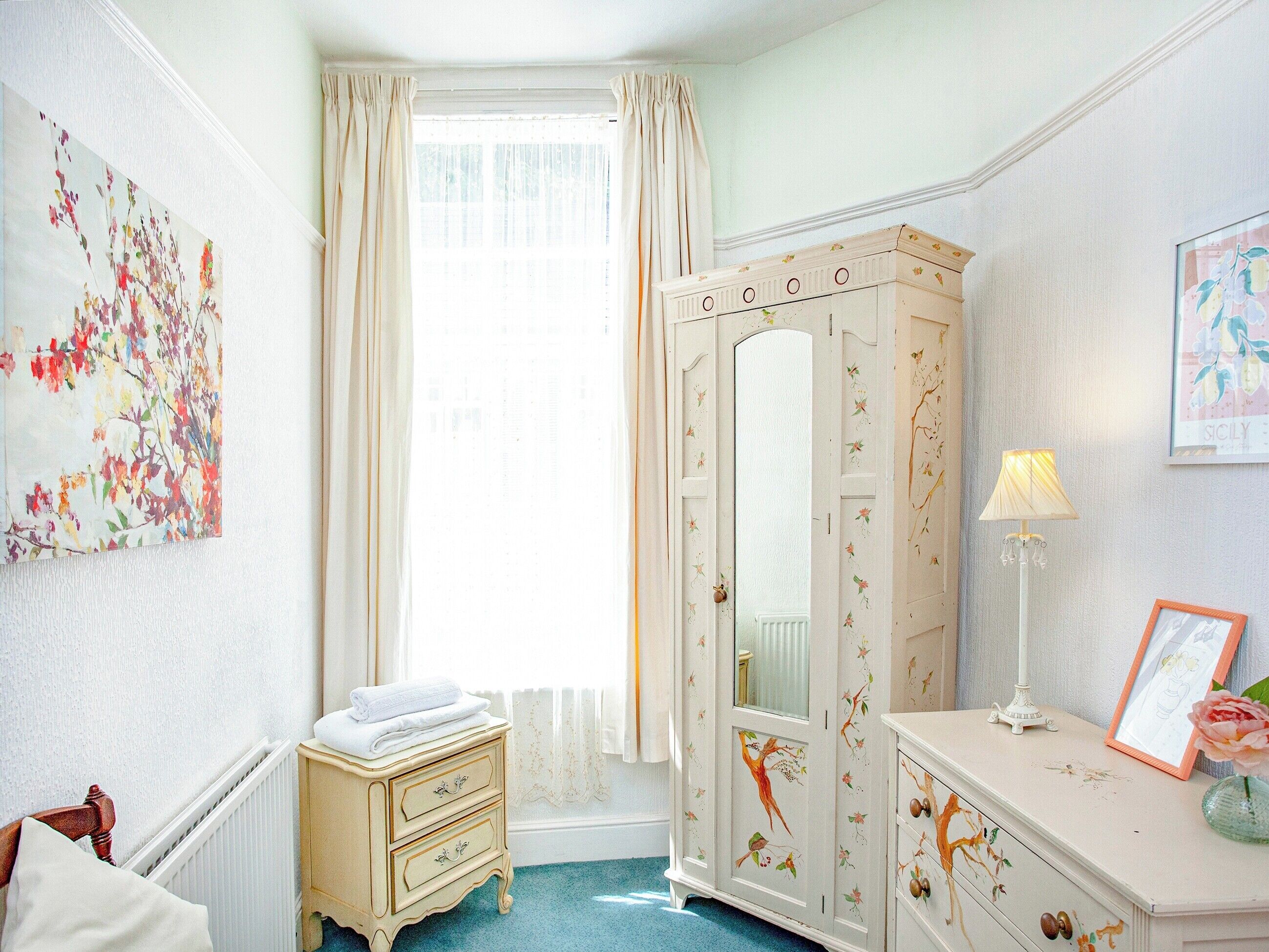 A room with a large window, a wardrobe with floral designs, a bedside table with a lamp, and a framed picture.