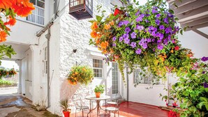 Un bâtiment blanc avec une terrasse aux carreaux rouges, une jardinière suspendue et une table avec des chaises.