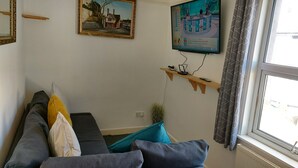 A living room with a sofa, a TV mounted on the wall, a window with curtains, and a small shelf with items. - Victoria Heights - Beautiful 3-bed in Gillingham K (Gillingham)