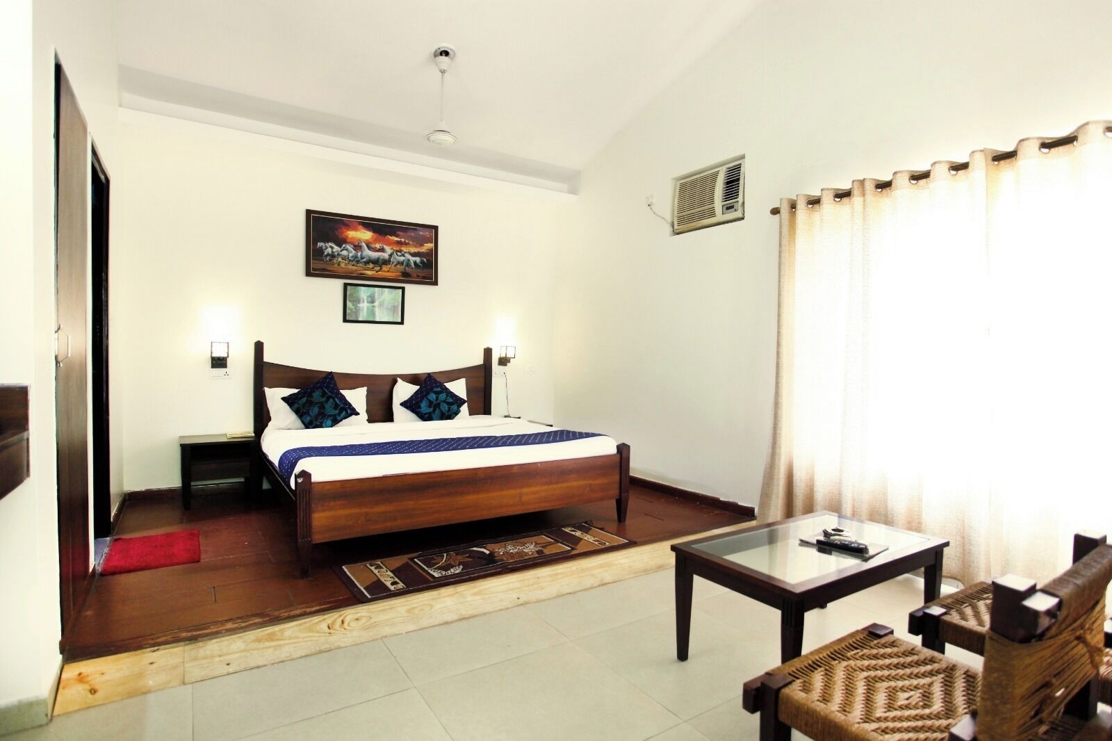 A hotel room with a wooden bed, a bedside table, a glass coffee table, and a woven chair.