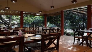 An outdoor dining area with wooden tables and chairs, surrounded by trees and a covered section with red pillars. - Corbett Aroma Park By Royal Collection (Ramnagar)
