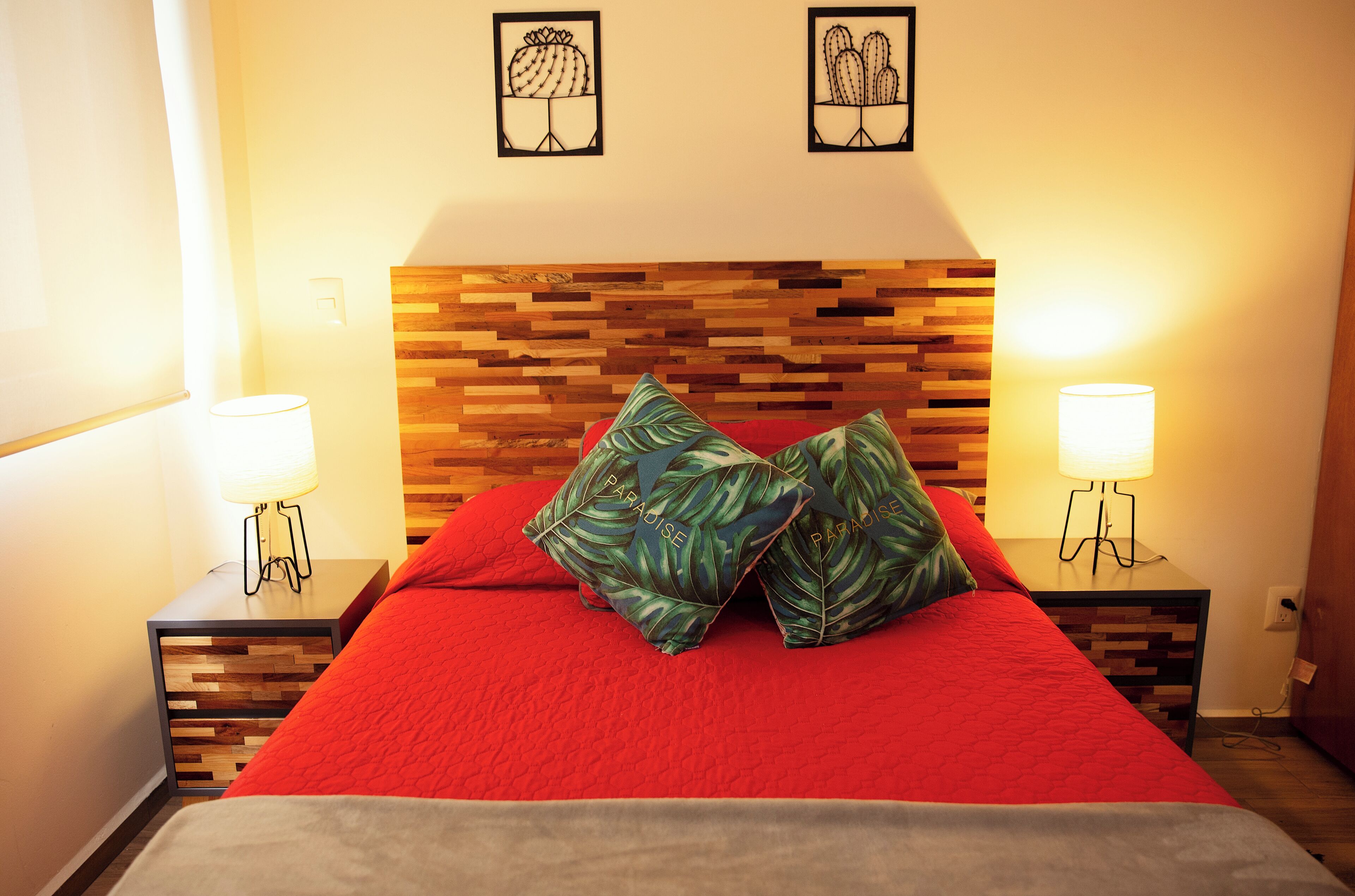 Een netjes opgemaakt bed met een rode deken en twee kussens, aan weerszijden met nachtkastjes voorzien van lampen, tegen een houten hoofdbord.