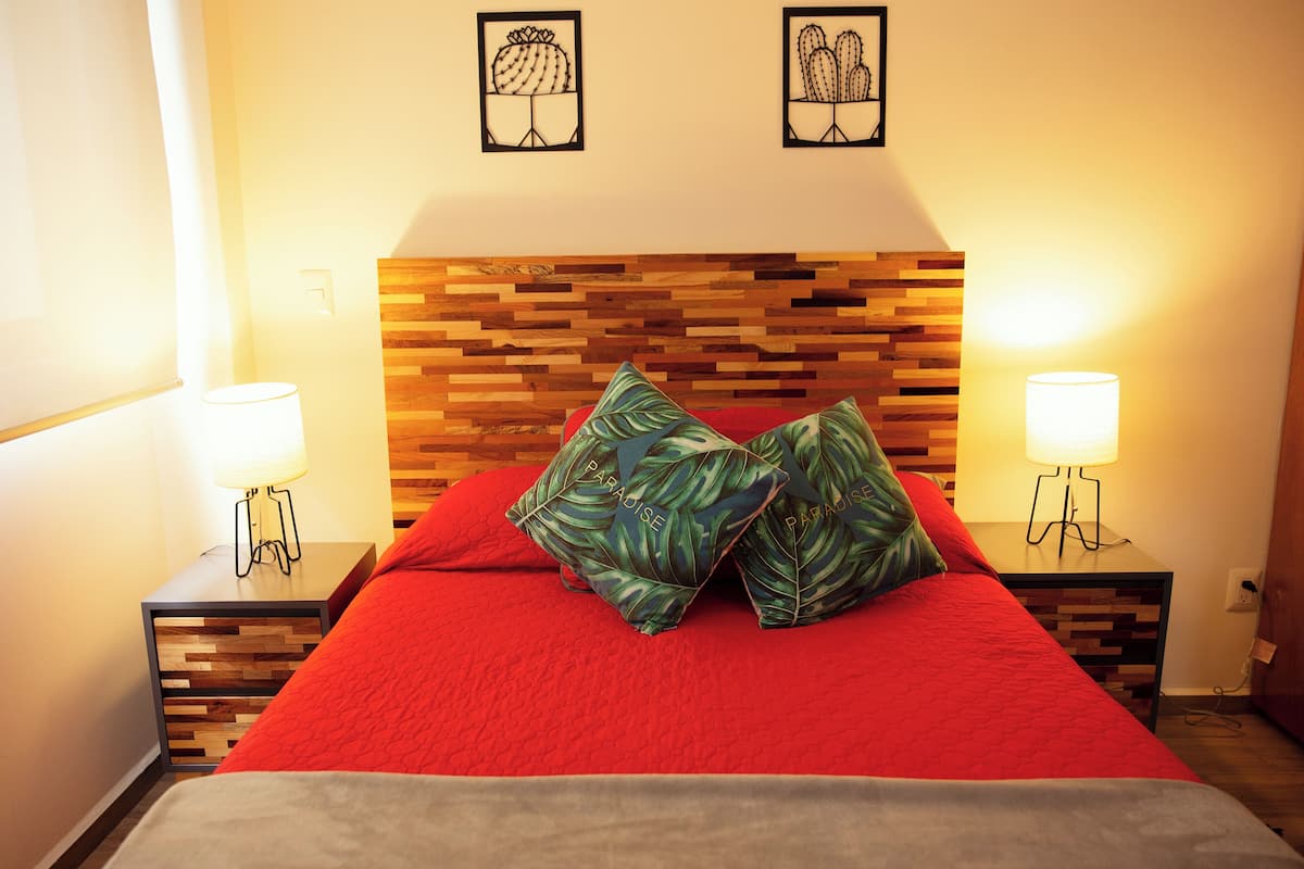 Una cama bien hecha con una colcha roja y dos almohadas, flanqueada por dos mesitas de noche con lámparas, frente a un cabecero de madera.