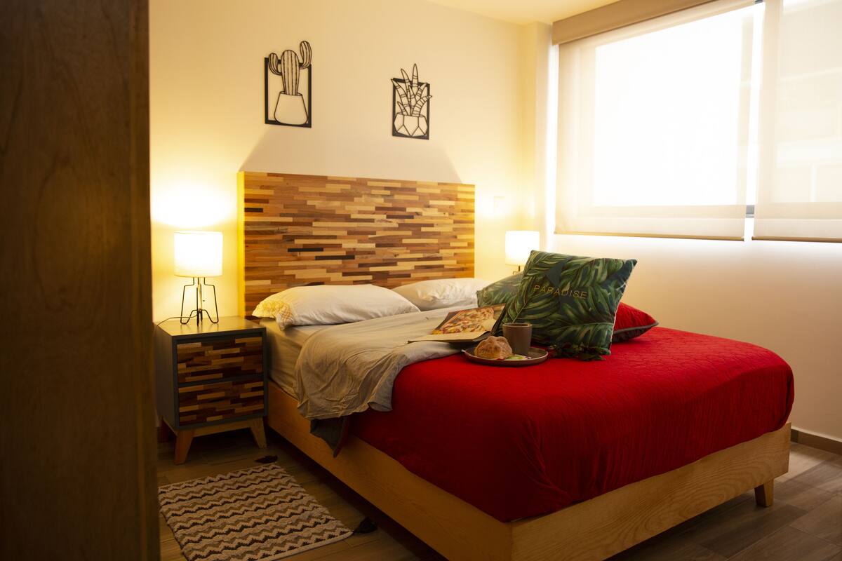 A neatly made bed with a red cover, a wooden headboard, and a bedside table with a lamp.