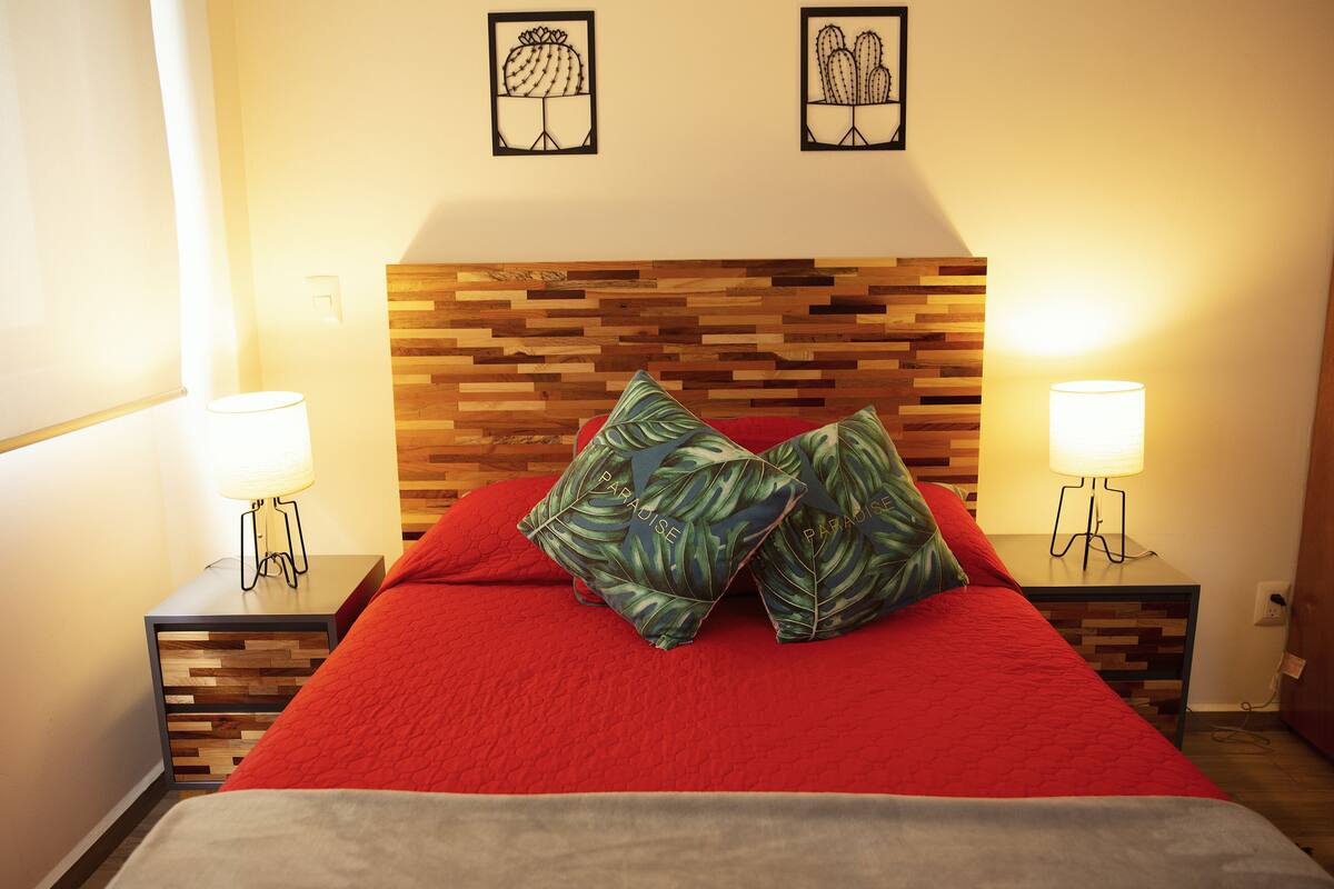 A neatly made bed with a red blanket and two pillows, flanked by two bedside tables with lamps, set against a wooden headboard.