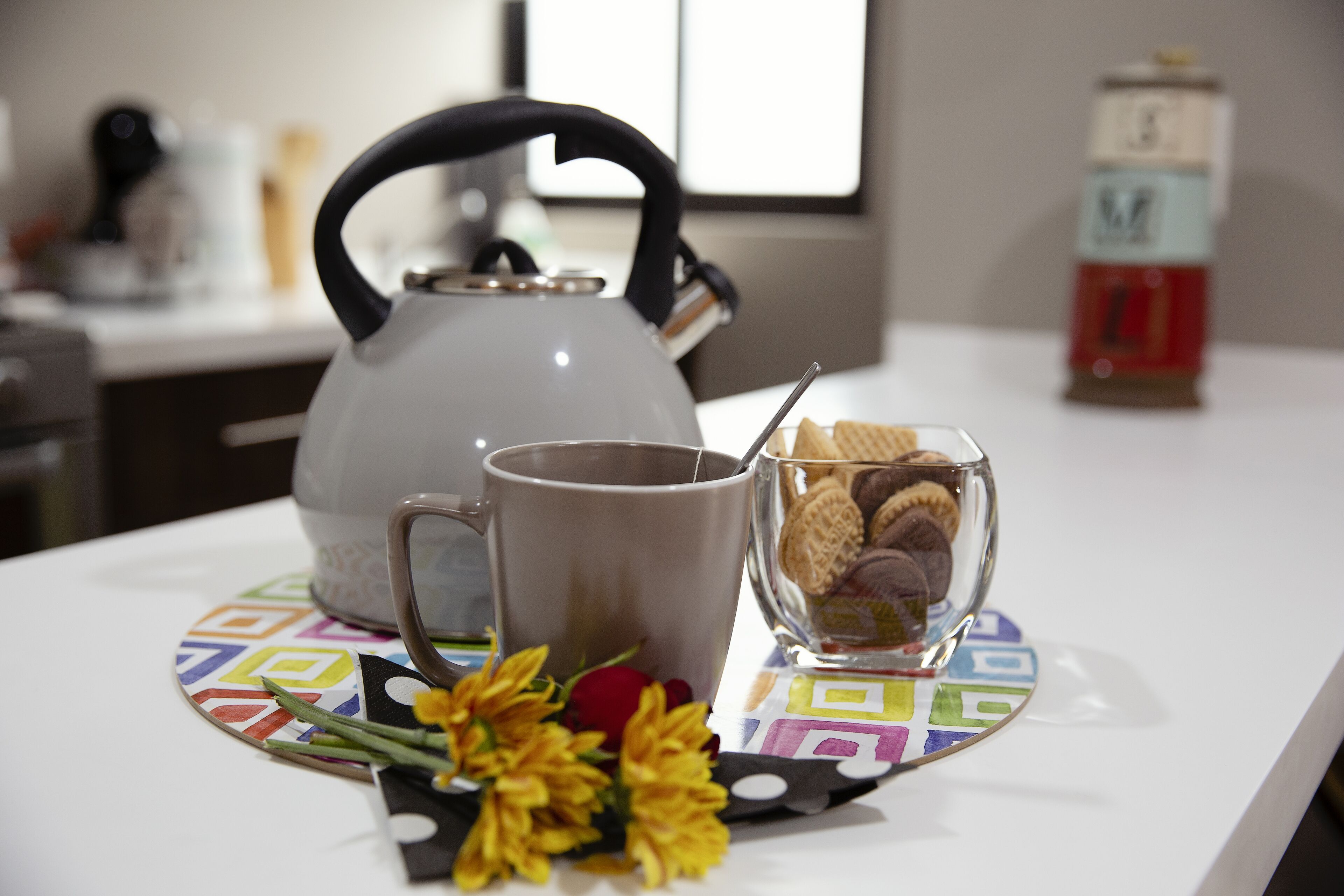 Een tafel met een waterkoker, beker, glas en koekjes, plus een kleurrijke onderzetter.