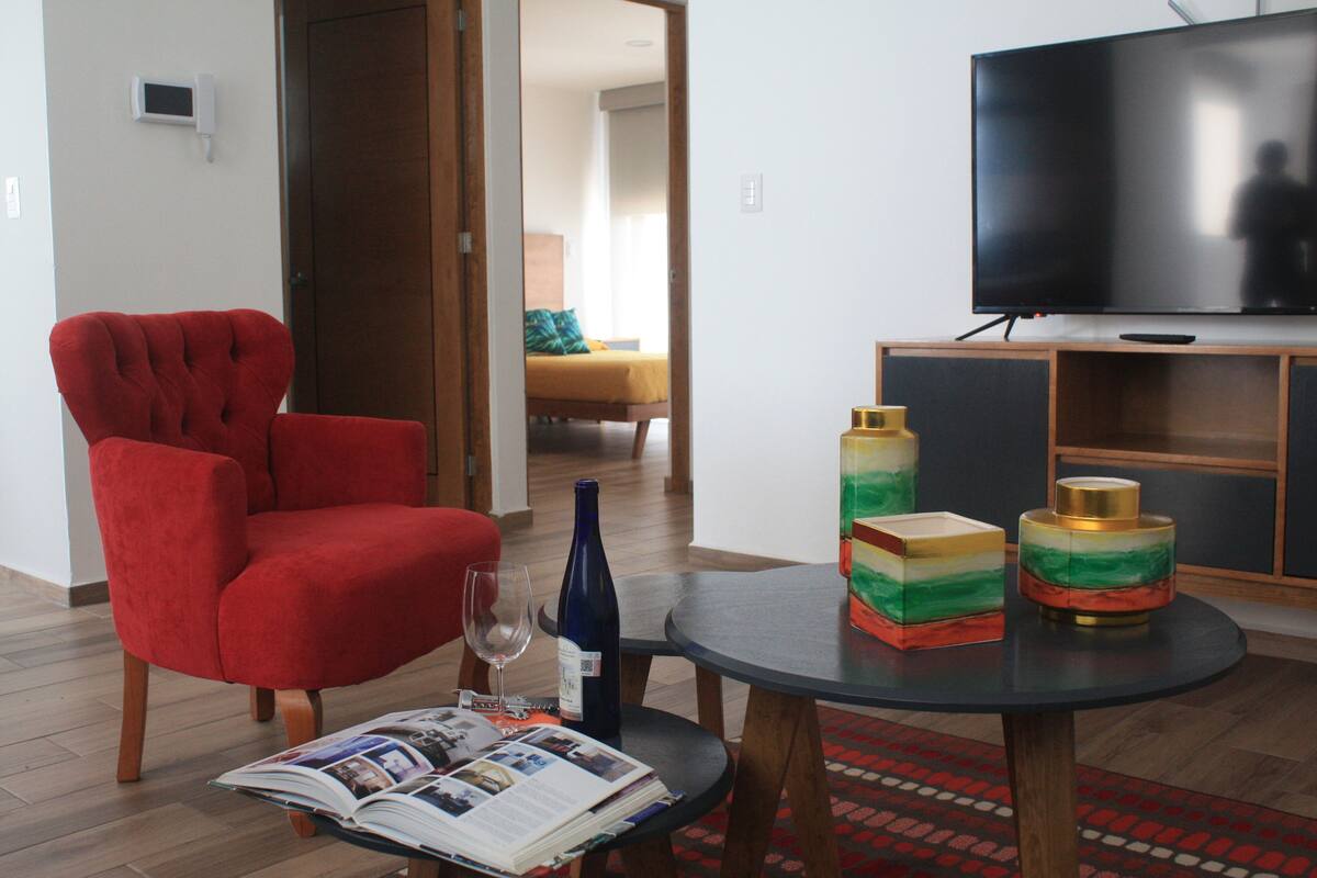 A living room with a red armchair, a round table with a magazine, a wine bottle, and decorative items.