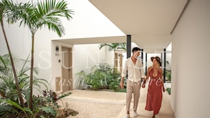A couple walking in a modern, well-lit hallway with a palm tree and plants, leading to a door labeled "107L". - Hunab Tulum (Tulum)