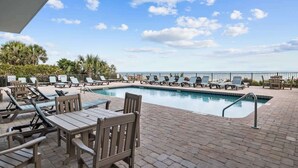A pool area with lounge chairs and tables, a clear sky, and a view of the ocean. - Renovated Condo with Gorgeous Ocean Views (Myrtle Beach)