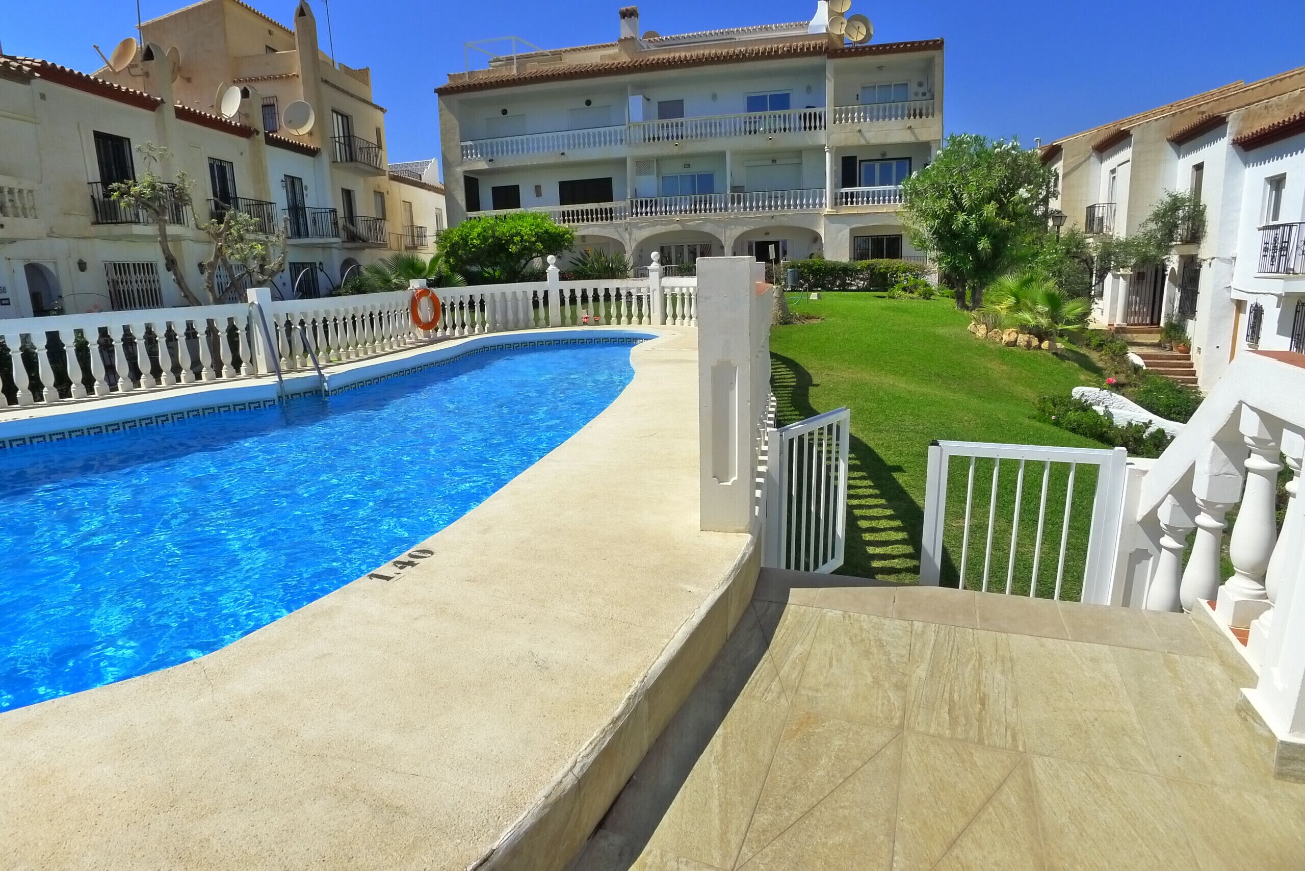 A swimming pool with a white fence, a clear blue sky, and a building with balconies in the background.