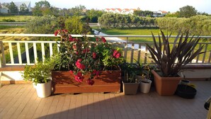 A balcony with a wooden bench, potted plants, and a view of a green landscape with trees and buildings.