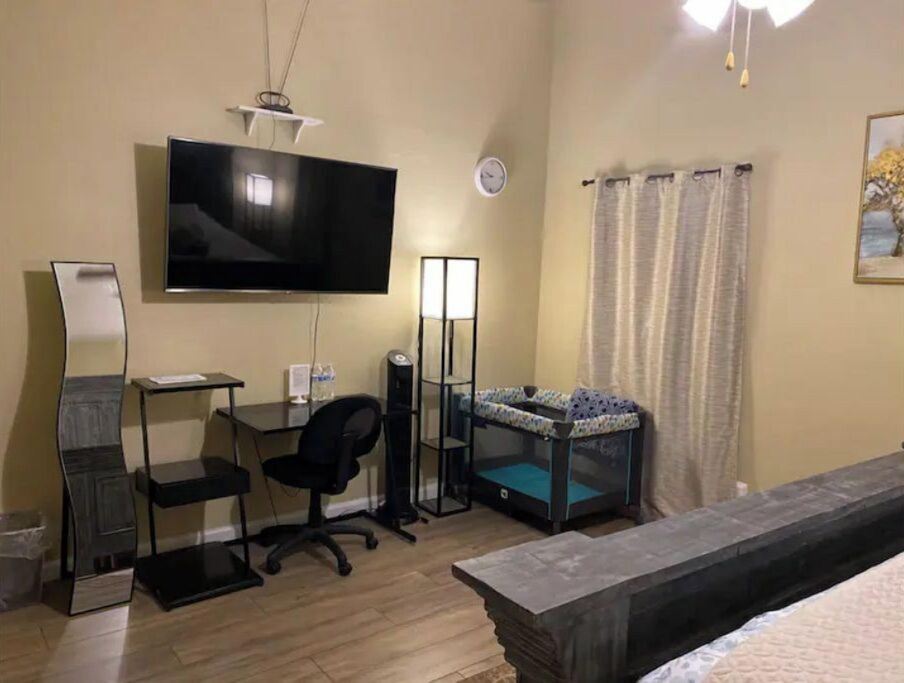 A hotel room with a bed, a desk with a chair, a television, a bedside table, a lamp, a clock, and a window with curtains.