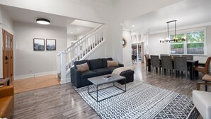A spacious living area with a dark-colored sofa, a coffee table, and a patterned rug. A staircase leads to another room, and a dining area with chairs and a table is visible.