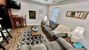 Uma sala de estar com um sofá cinzento, uma mesa de centro, uma televisão na parede e um letreiro com a palavra 'HOME'.