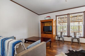 Ein Wohnzimmer mit einer Couch, einer Holzkommode mit Fernseher, zwei Stühlen und einem Fenster mit Jalousien.