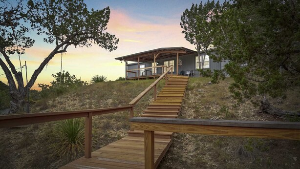 A wooden staircase leading to a modern house with a covered porch, surrounded by trees and dry grass. - Hondo Creek Hideaway (Tarpley)