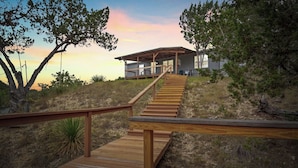 En trætrap fører op til et moderne hus med en overdækket veranda, omgivet af naturlig beplantning.