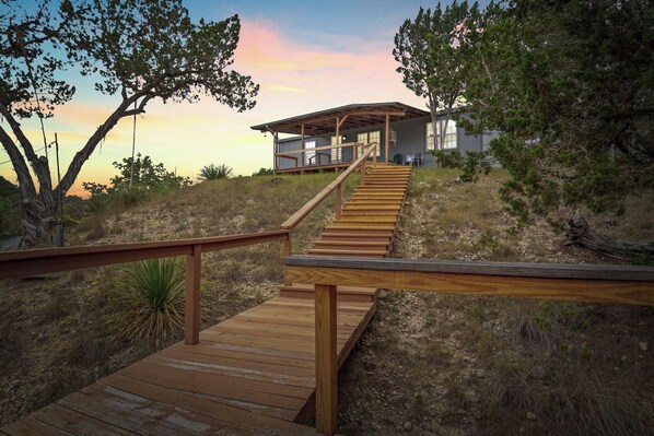 A wooden staircase leading to a modern house with a covered porch, surrounded by natural vegetation.