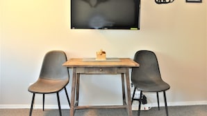 A small room with a TV mounted on the wall, a wooden table with two chairs, and a power outlet on the floor. - Whitefoot Lodge Studio in Village Centre (Beaverdell)