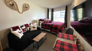 A cozy room with a black sofa, a wooden bench, a bed with red and black bedding, and a TV on the wall. - Whitefoot Lodge Studio in Village Centre (Beaverdell)