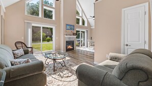A spacious living room with a fireplace, large windows, and a ceiling fan.