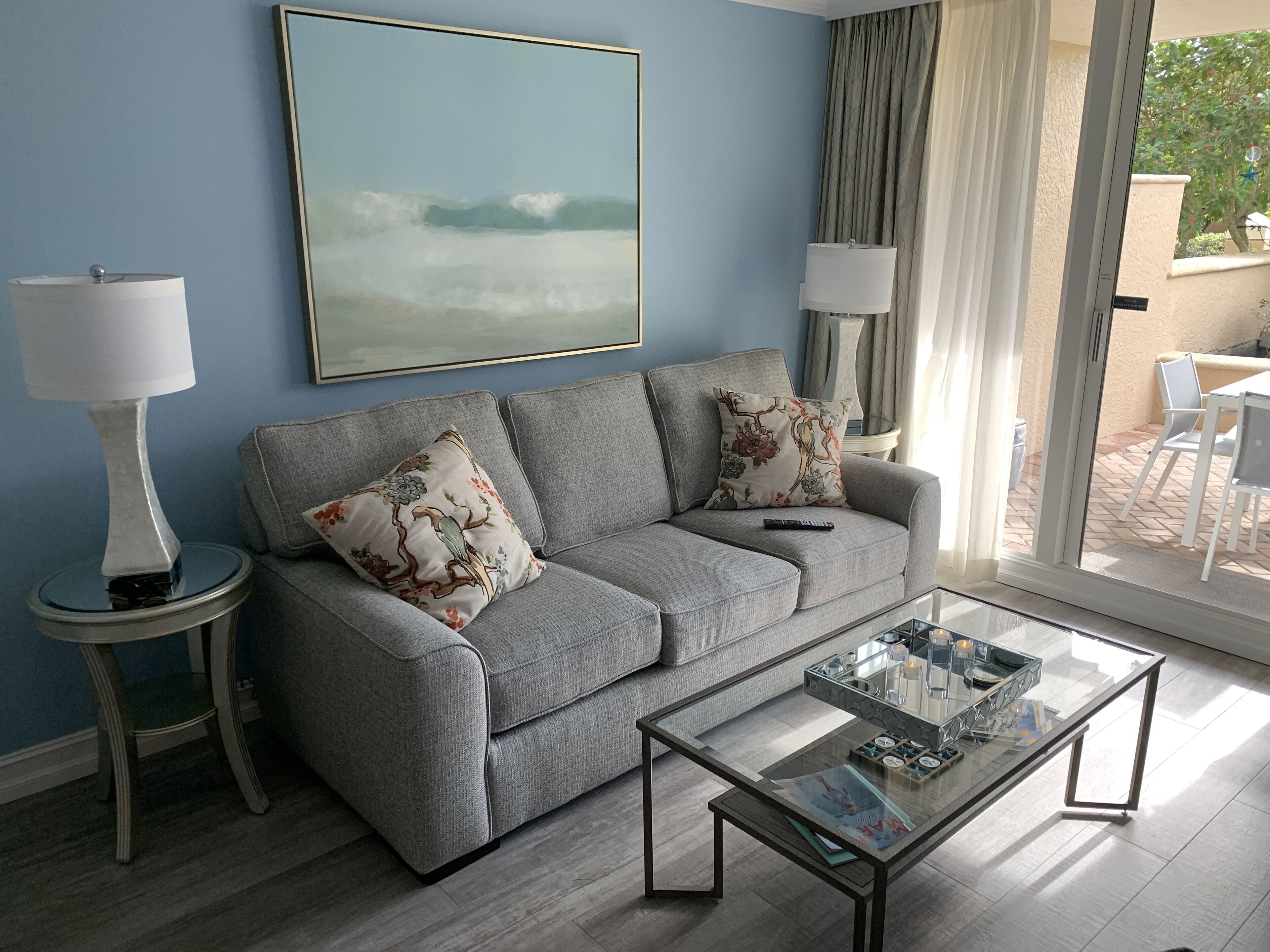 Una sala de estar moderna con un sofá gris, dos cojines decorativos, una mesa de centro de vidrio y una mesita de noche con una lámpara.