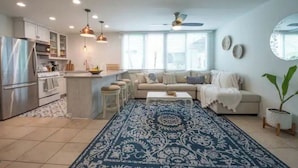 A modern kitchen with a central island, a dining area with a sofa and coffee table, and a living room with a large window and a ceiling fan. - Cottage by the Sea steps to beach with heated pool (Carpinteria)