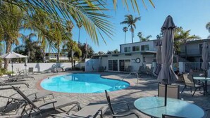 A pool area with lounge chairs and umbrellas, surrounded by palm trees and a modern building. - Cottage by the Sea steps to beach with heated pool (Carpinteria)